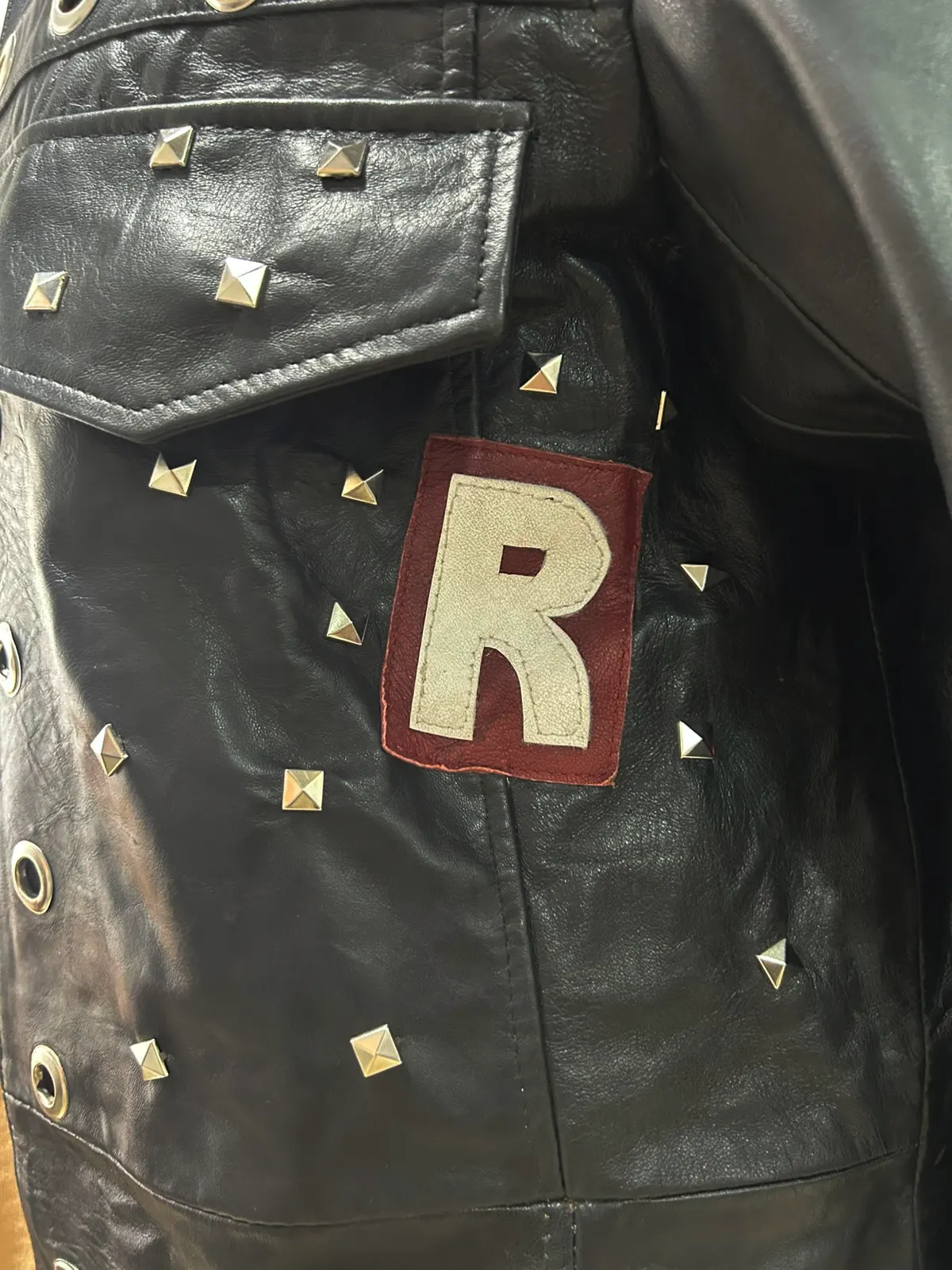 Close-up detail of the WWE Edge Royal Rumble Trench Coat showing a white capital letter "R" patch sewn onto a red leather background surrounded by square metal studs.