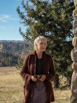 Helen Mirren as Cara Dutton wearing a long brown knit coat cardigan and leather gloves in a rural field