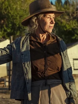 Helen Mirren as Cara Dutton in "1923," wearing a wide-brimmed brown hat, a dark brown button-up shirt, and an open grey jacket, looking thoughtfully off-camera in warm evening sunlight.