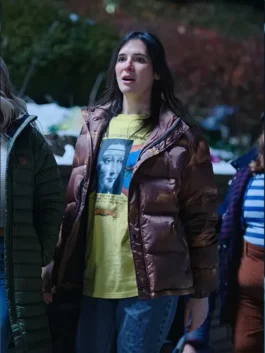 A surprised young woman wearing a metallic brown puffer jacket and a yellow graphic t-shirt outdoors at night