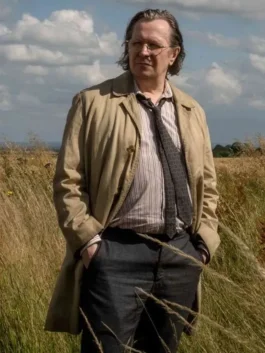 Full-length shot of Gary Oldman portraying Jackson Lamb in an outdoor field setting. He is wearing his iconic Jackson Lamb beige trench coat and dark trousers, emphasizing the Gary Oldman Jackson Lamb coat from the spy series.