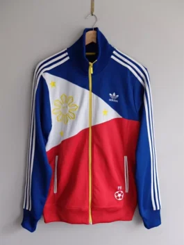 An Adidas jacket Philippines edition hanging on a wall, featuring the Filipino flag sun and stars embroidery and a white soccer ball PH logo.
