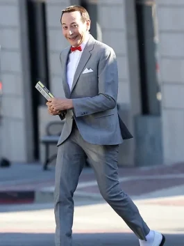Pee Wee Herman smiling in his signature grey pee wee herman outfit with a red bow tie and white shoes while holding a booklet.