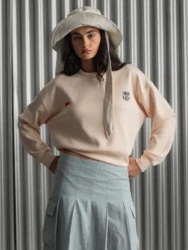 Woman wearing a peach Barça Essentials Crew Sweatshirt with FC Barcelona crest, styled with a light blue skirt and wide-brim hat