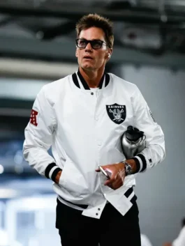 Tom Brady wearing a white Raiders varsity jacket with the team's shield logo patch, walking and holding a water bottle.