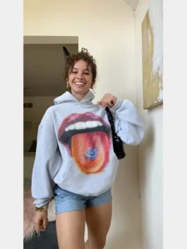 A woman smiling while wearing a grey homesick hoodie from the homesick apparel line, featuring a vibrant mouth and tongue graphic.