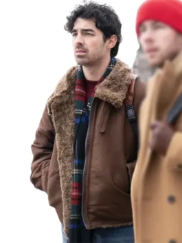 Joe Jonas in a rustic brown shearling-lined suede leather bomber jacket with a furry collar, a dark red sweater underneath, and a plaid green, blue, and red scarf. He is looking off to the side, standing next to a blurred figure wearing a red beanie and a tan coat.