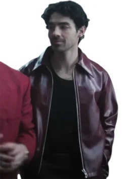 Joe Jonas in a glossy, zippered maroon leather jacket with a wide collar, worn over a black t-shirt. He is looking off to the side, and a person in a red blazer is partially visible on the left.