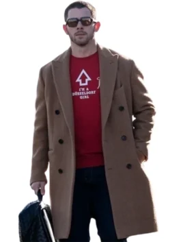 Nick Jonas sitting down, wearing a camel-colored coat over a dark green button-down shirt, dark pants, and a bright red knit beanie. He is looking off to the side, with his arms crossed and some blurred greenery in the foreground.