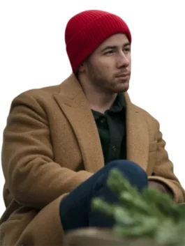 Nick Jonas standing in a long, camel-colored double-breasted coat and dark jeans. He wears dark sunglasses and a red sweatshirt underneath that reads "I'M A DÜSSELDORF GIRL" with an arrow pointing up. He is holding a black bag in his left hand.