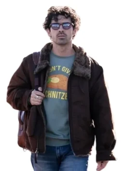 Joe Jonas, wearing sunglasses and a dark brown bomber jacket with a shearling collar, carries a brown leather backpack over his shoulder. He's wearing a light green graphic t-shirt that says "I DON'T GIVE A SCHNITZE" and blue jeans.