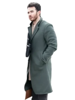 Kevin Jonas standing and looking forward, wearing a dark green coat with a zipper closure and a dark knit beanie on his head.
