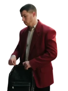 Nick Jonas, in a deep red blazer and a light-colored, slightly distressed shirt, leans over to adjust a black backpack he is holding. His hair is closely cropped.