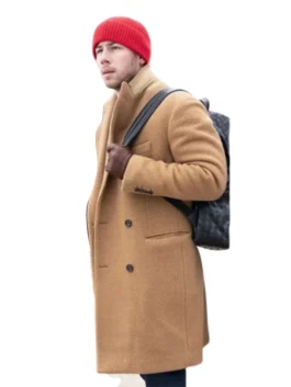 Nick Jonas is seen in profile wearing a tan wool overcoat with a lighter brown collar, a red knit beanie, and brown leather gloves. He has a black quilted backpack slung over his shoulder.