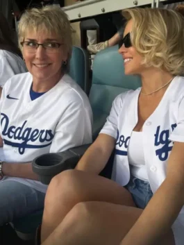 Sydney Sweeney smiling while seated at a Dodgers game next to a woman her friend, both wearing Sydney Sweeney Dodgers white shirt jerseys. Sydney Sweeney is wearing a white LA Dodgers Nike jersey open over a white top. This showcases the Sydney Sweeney Dodgers game outfit.