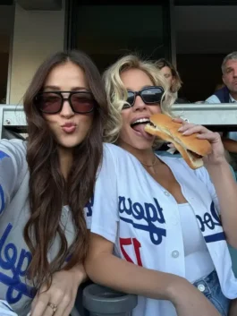 Sydney Sweeney and a friend posing with a playful expression while seated at a Dodgers game. Sydney Sweeney is licking a hotdog and wearing a white Sydney Sweeney Dodgers white shirt jersey open over a white top, highlighting the Sydney Sweeney Dodgers appearance and Sydney Sweeney MLB jersey.