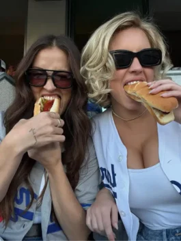 Sydney Sweeney and a friend at a Dodgers game, both wearing Dodgers jerseys and sunglasses, eating hotdogs. Sydney Sweeney is wearing a Sydney Sweeney white Dodgers jersey and is shown with a hotdog near her mouth, highlighting the Sydney Sweeney Dodgers game look and Sydney Sweeney LA Dodgers jersey.