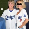 Sydney Sweeney posing with a woman in front of a Dodgers backdrop. Sydney Sweeney is wearing a Sydney Sweeney white Dodgers jersey open over a white top, paired with denim shorts, featuring the 'Dodgers' script and the number '17' for Shohei Ohtani. This captures her Sydney Sweeney Dodgers fan style.