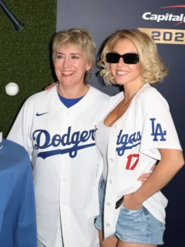 Sydney Sweeney posing with a woman in front of a Dodgers backdrop. Sydney Sweeney is wearing a Sydney Sweeney white Dodgers jersey open over a white top, paired with denim shorts, featuring the 'Dodgers' script and the number '17' for Shohei Ohtani. This captures her Sydney Sweeney Dodgers fan style.