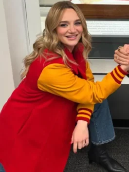 Alana Higman is sitting down and smiling, showing her long red coat with yellow varsity-style sleeves, wide-leg blue jeans, and black ankle boots. The coat's cuffs have red and yellow stripes.