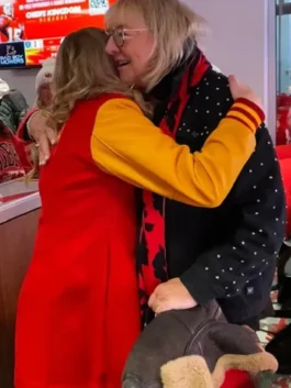 Alana Higman, wearing a red coat with yellow sleeves, is embracing an older blonde woman who is wearing glasses, a black jacket with white dots, and a red and black patterned scarf.