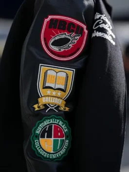 A close-up view of the sleeve of the black leather varsity jacket, showcasing several embroidered patches. Patches include the NFL Shield, a logo referencing 