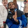 NFL player Damar Hamlin smiles while posing with a young girl. He is wearing a blue and black puffer jacket with a ribbed collar and an NFL shield logo, and he is holding up a peace sign.