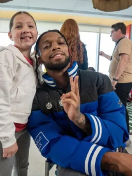 NFL player Damar Hamlin smiles while posing with a young girl. He is wearing a blue and black puffer jacket with a ribbed collar and an NFL shield logo, and he is holding up a peace sign.
