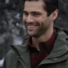 Close-up of actor Matthew Daddario smiling outdoors. He has dark hair, a beard, and is wearing an olive green hooded jacket over a red and black plaid shirt.