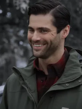 Close-up of actor Matthew Daddario smiling outdoors. He has dark hair, a beard, and is wearing an olive green hooded jacket over a red and black plaid shirt.