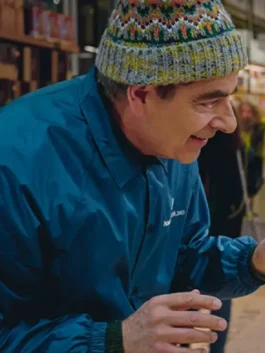 Close-up of Rowan Atkinson smiling in a brightly colored knit beanie and the 'Housesitters Deluxe' blue jacket, suggesting a costume or jacket available for sale.