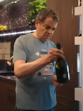 Close-up of Rowan Atkinson as Trevor Bingley in the comedy series Man vs Baby 2025, wearing a light blue/grey, round-neck Fish Printed T-Shirt while holding a bottle of sparkling wine. The T-Shirt features a horizontal print of stylized fish.