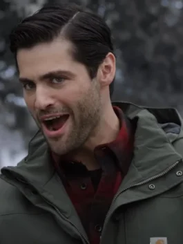 Close-up of actor Matthew Daddario looking surprised or excited, with his mouth open. He is wearing an olive green hooded work jacket with a small brand logo on the chest, layered over a red plaid shirt.