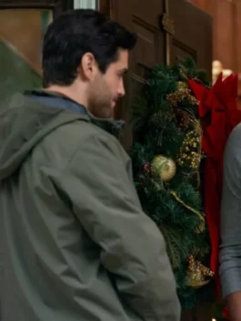 Actor Matthew Daddario, viewed from the back and side, is standing by a dark wooden door decorated with a large green Christmas wreath featuring gold accents and a red bow. He is wearing an olive green hooded jacket.