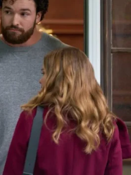 Holland Roden, with long wavy strawberry-blonde hair, is seen from the back wearing a burgundy or deep red coat and a shoulder bag. A tall man with a full beard and a gray sweater is standing behind her, looking over her shoulder.