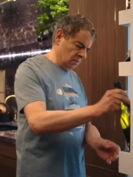 Mid-shot of Rowan Atkinson looking concerned while pulling a bottle from a wine rack. He is wearing a light blue/grey, crew-neck Fish Printed T-Shirt which features a graphic of stylized fish. This image highlights the casual Rowan Atkinson T-Shirt outfit.