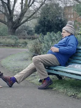 Rowan Atkinson sitting on a park bench, looking distressed, wearing a knit beanie, a blue jacket, khaki trousers, and polka-dot socks with brown shoes. This is from the 