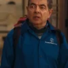 Close-up of Rowan Atkinson from "Man vs Baby," wearing the distinctive Housesitters Deluxe Blue Jacket with the logo visible. He is wearing a backpack and a patterned sweater underneath the Rowan Atkinson blue jacket for men.
