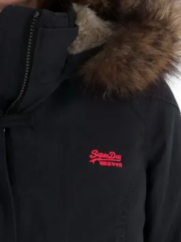 A close-up shot of the shoulder and collar of the black Superdry Study Duffle Jacket. It clearly shows the zipper detail, the textured brown Vegan fur hood trim, and the small, bright red embroidered Superdry logo on the chest.
