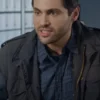 Close-up of actor Matthew Daddario looking sternly off-camera. He has dark hair and a slight beard, and is wearing a dark, possibly black or charcoal, field jacket over a blue plaid button-down shirt.