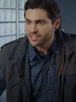 Close-up of actor Matthew Daddario looking sternly off-camera. He has dark hair and a slight beard, and is wearing a dark, possibly black or charcoal, field jacket over a blue plaid button-down shirt.