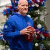 Sean McDermott, the bald-headed coach of the Buffalo Bills, stands in front of a Christmas tree decorated with red, white, and blue ornaments and lights. He is wearing a bright blue Buffalo Bills hooded sweatshirt with the team logo on the chest and an NFL shield patch on the sleeve, holding a football with both hands.