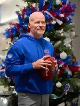 Sean McDermott, the bald-headed coach of the Buffalo Bills, stands in front of a Christmas tree decorated with red, white, and blue ornaments and lights. He is wearing a bright blue Buffalo Bills hooded sweatshirt with the team logo on the chest and an NFL shield patch on the sleeve, holding a football with both hands.