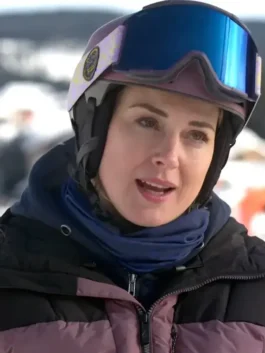Woman wearing a ski helmet and goggles with a black and mauve puffer jacket and blue neck warmer outdoors.