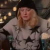Blonde woman wearing a white star sweater and black beanie in a festive outdoor setting with string lights.