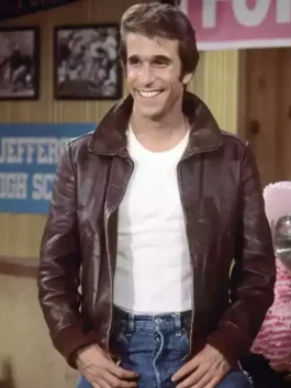 Henry Winkler as Fonzie wearing his signature brown leather jacket over a white t-shirt and blue jeans, standing and smiling on the Happy Days set.