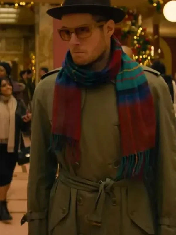 Connor Swindells wearing a dark hat, tinted glasses, a green trench coat, and a multicolored scarf walks through a festively decorated indoor space.