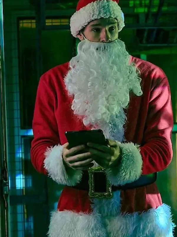 Connor Swindells wearing a red Santa coat with white fur trim and beard, holding a tablet in a festive indoor setting.