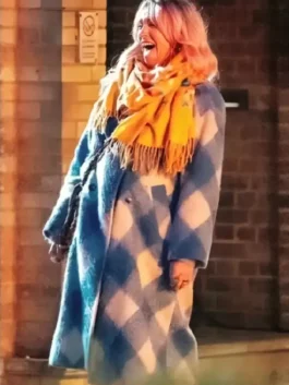 Woman wearing a blue and white check long coat with a mustard yellow scarf, standing outdoors near a brick wall in warm light.