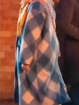 Side view of a woman wearing a blue and white check long coat with a mustard scarf, walking outdoors near a brick wall.